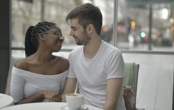 egy fekete hajú nő és egy fehér pólós férfi egy kávézó asztalánál ülnek randevún