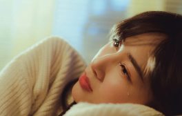 A woman in a sweater lying down and shedding a tear in a warm setting.